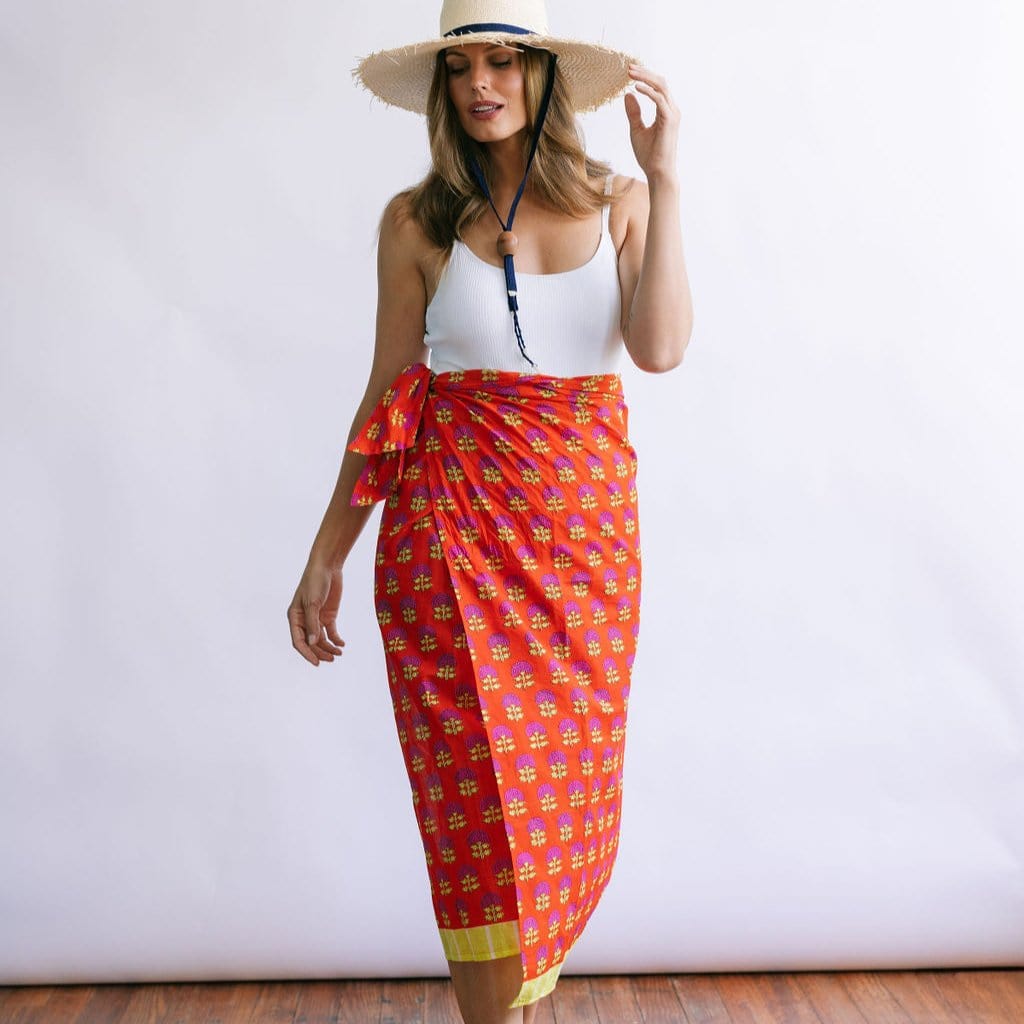 A woman wearing a white tank top and a wide-brimmed hat, draped in a red and orange sarong with a geometric pattern.