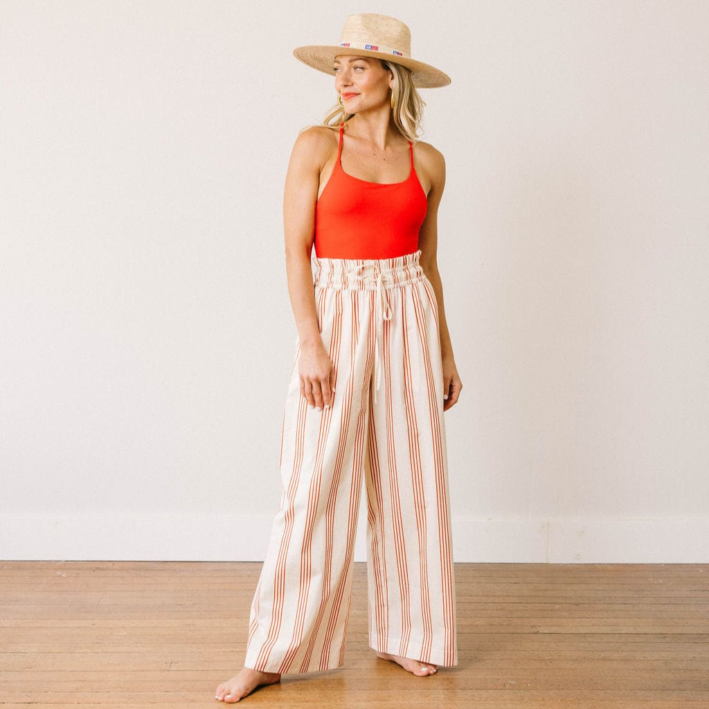 Woman wearing a red tank top and striped pants with a straw hat on a plain background
