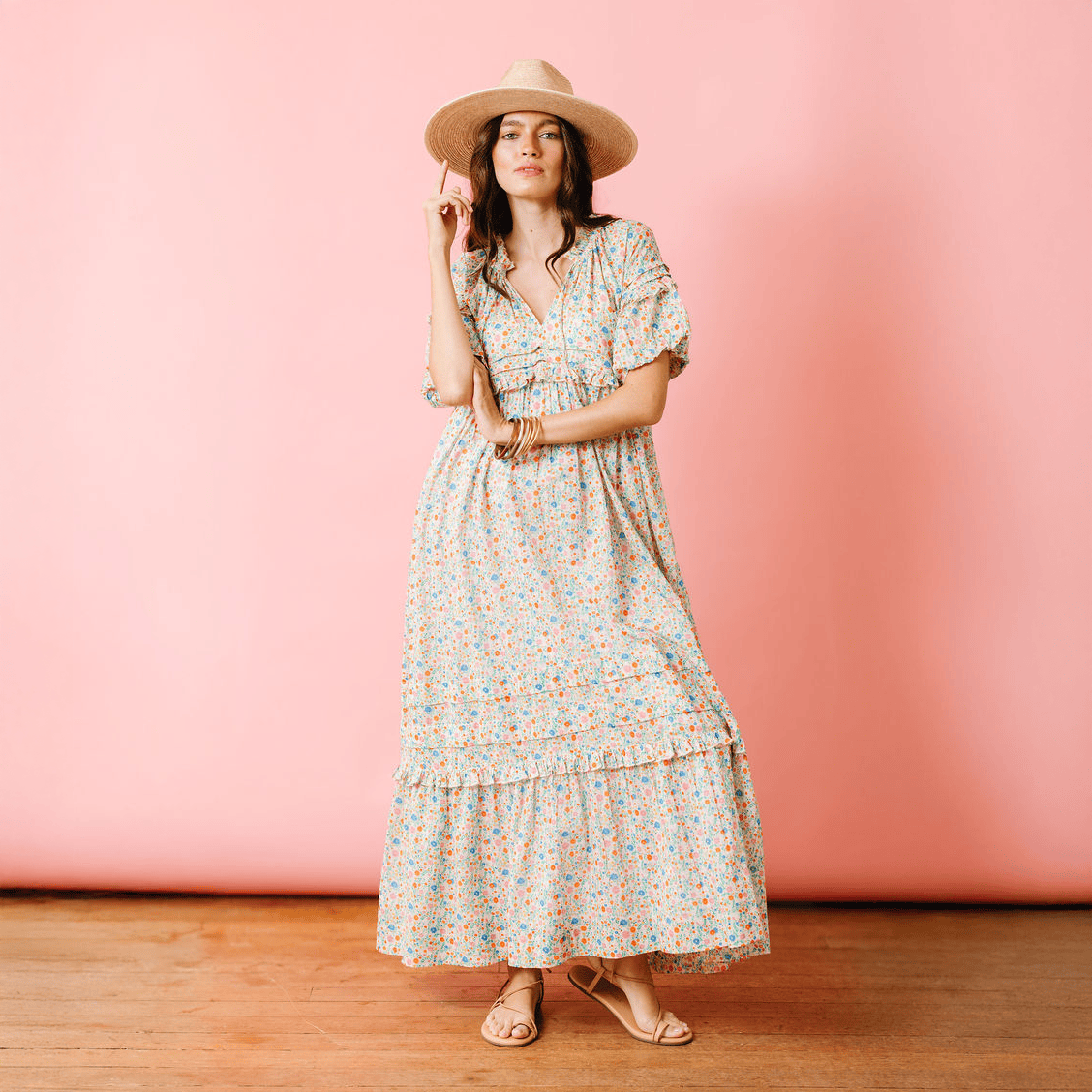Woman in a floral dress and hat standing against a pink wall.