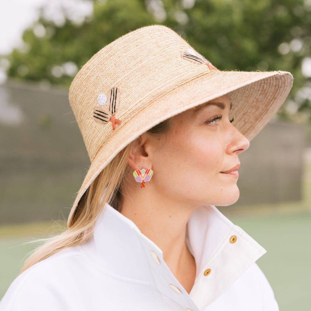 A person wearing a straw hat and a white jacket, looking to the side. The person is also wearing colorful pickleball stud earrings with stripes and paddle and ball details.