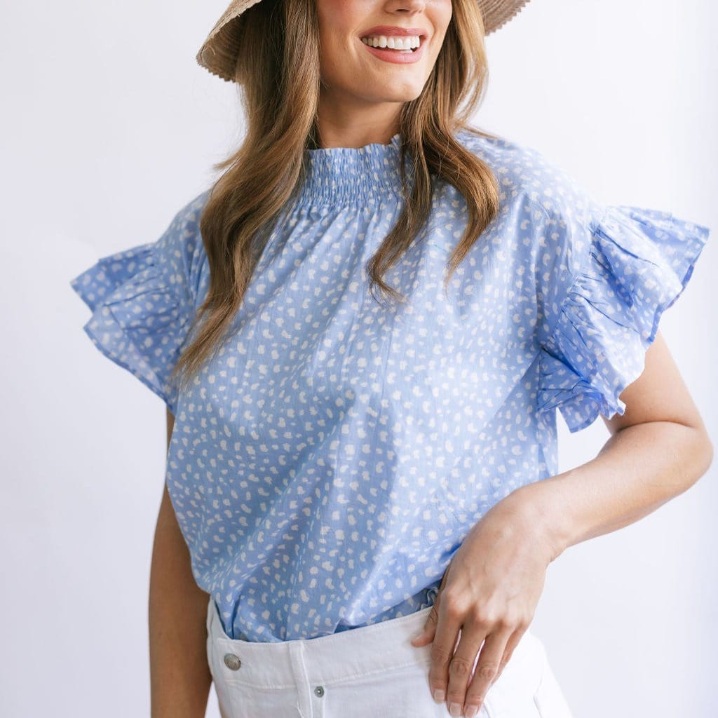 A woman wearing a light blue dot flutter top with ruffle sleeves and a ruched neckline, paired with white pants.
