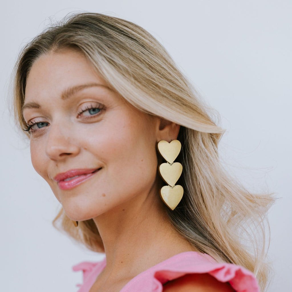 A woman wearing gold metallic heart earrings with a pink top.