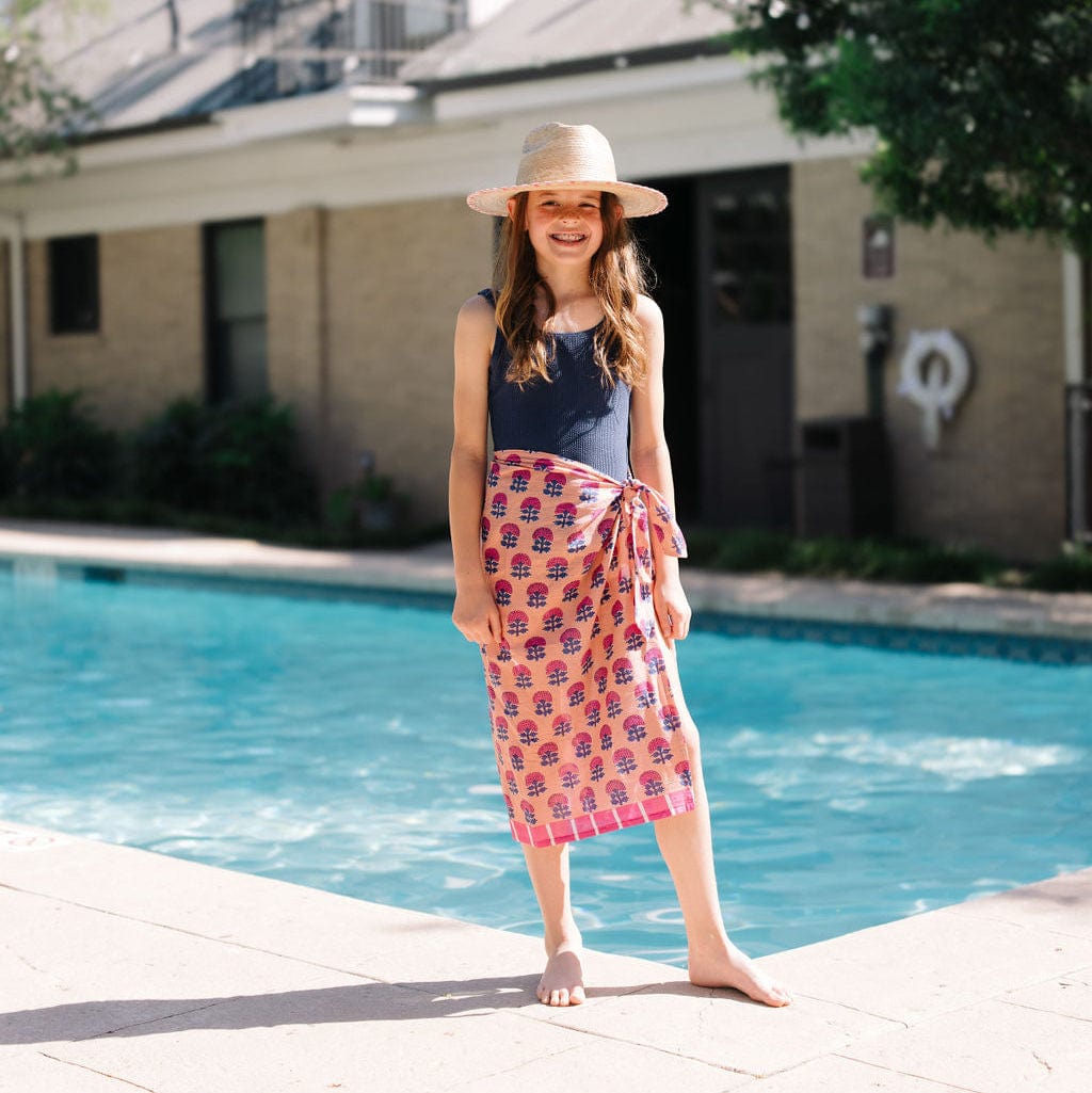 Young girl standing by a pool wearing a blue top and patterned pink skirt.