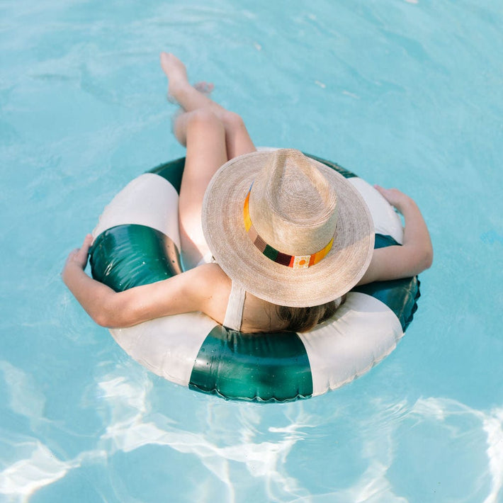 Person wearing a straw hat and sunglasses on a green and white inflatable ring in clear blue water.