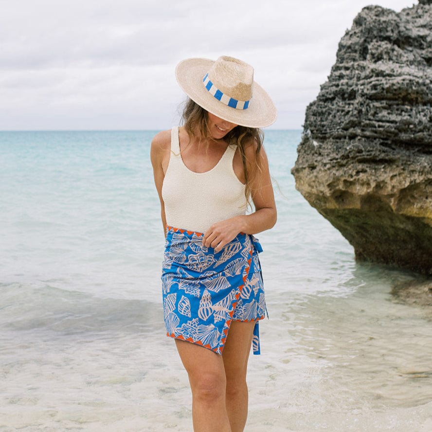 Woman in a white tank top and blue patterned skirt standing on a beach with a large rock formation in the background.