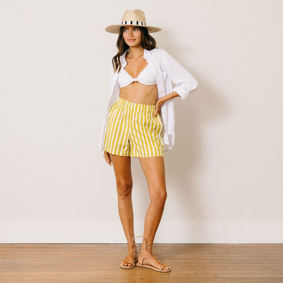 Woman wearing a white top, yellow striped shorts, and a straw hat against a plain background