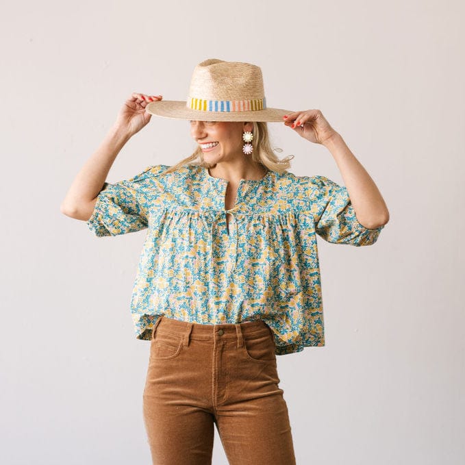 Woman wearing a floral blouse and brown pants, holding a straw hat with colorful band.