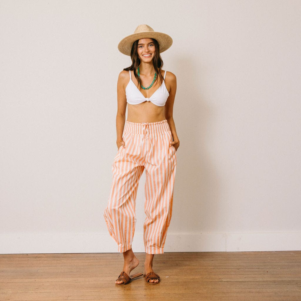 Woman wearing a white bikini top and striped orange pants with a straw hat against a plain background