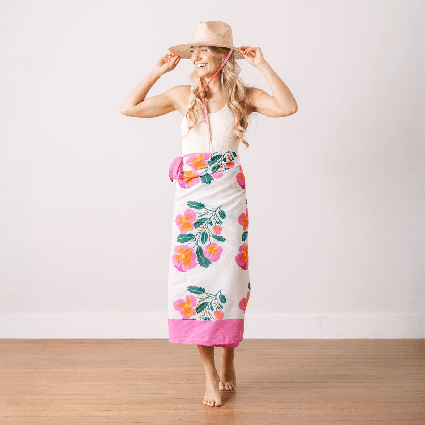 A woman standing in a room with a white background, wearing a white top and a pink flower sarong, with a hat in her hands.