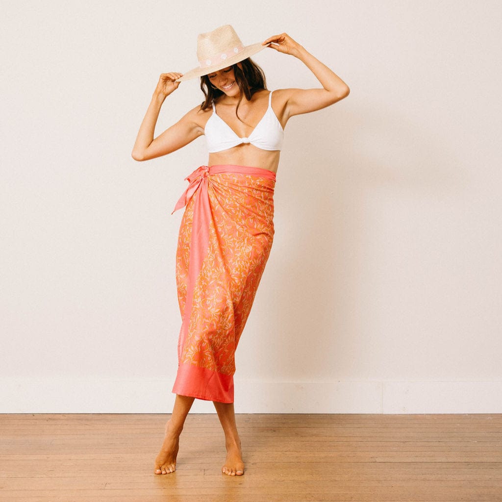 Woman wearing a white bikini top and orange patterned sarong against a plain background