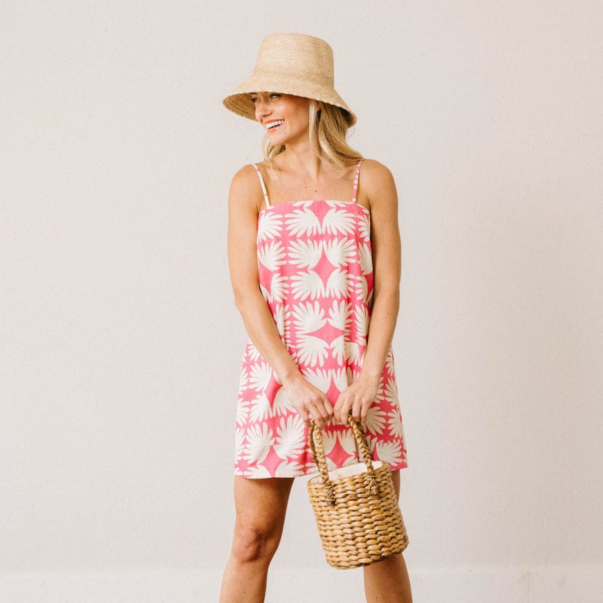 Woman wearing a pink dress with white floral pattern, holding a straw bag, against a plain background