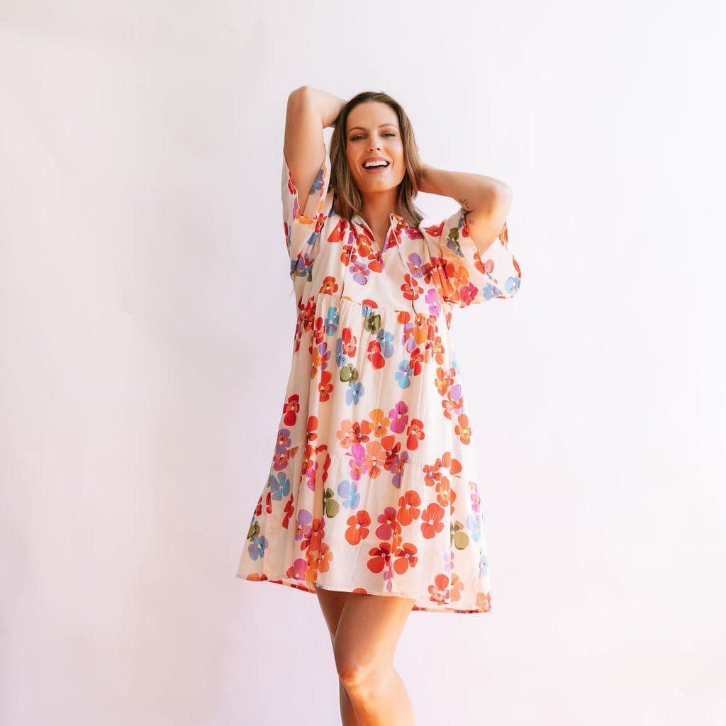 A woman wearing a floral dress with a V-neck and 3/4 sleeves standing with her hands raised and elbows out, against a white background.