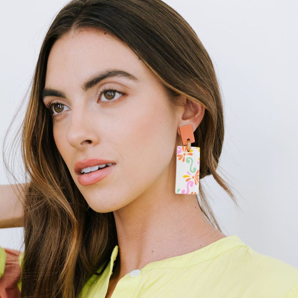A woman wearing a yellow top and showcasing earrings with a geometric pattern in multiple colors including orange, green, pink, yellow, and white.