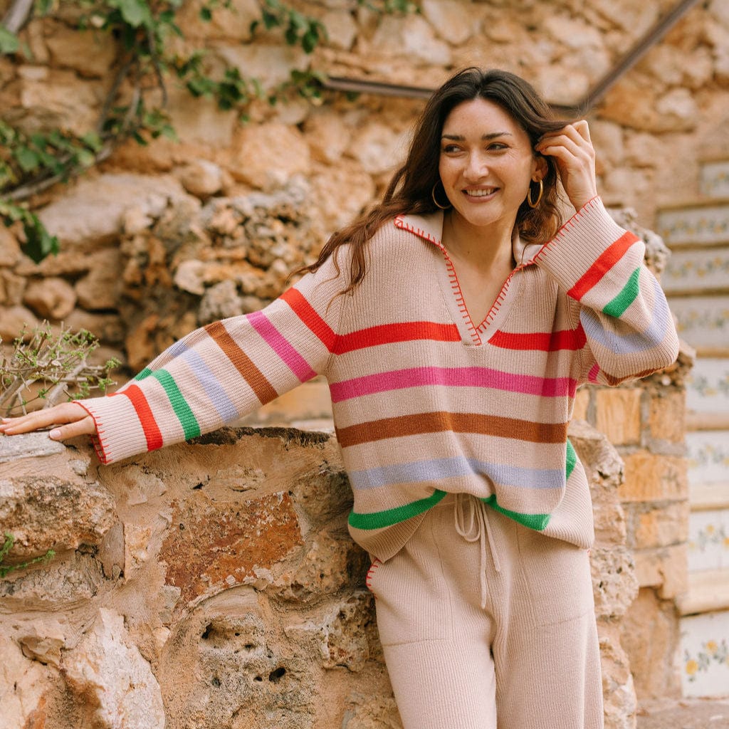 Woman wearing a colorful striped sweater and beige pants standing against a stone wall.