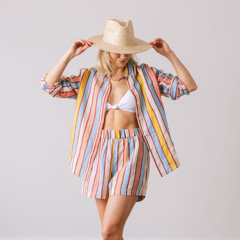 A woman modeling a Seaplane Stripe Bermuda Set, consisting of a button-down shirt and matching shorts with a striped pattern. She is also wearing a wide-brimmed hat.