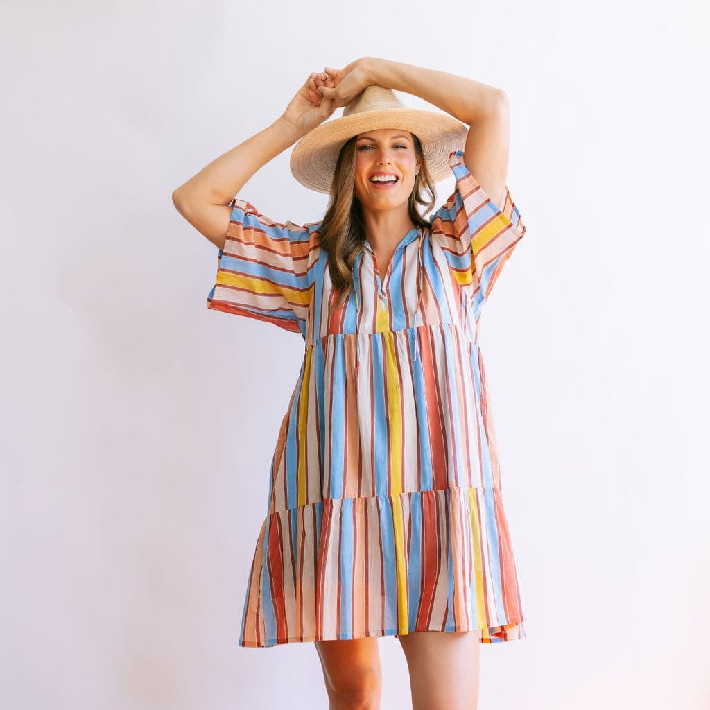 A woman wearing a multicolored striped knee-length dress with sleeves, paired with a wide-brimmed hat.