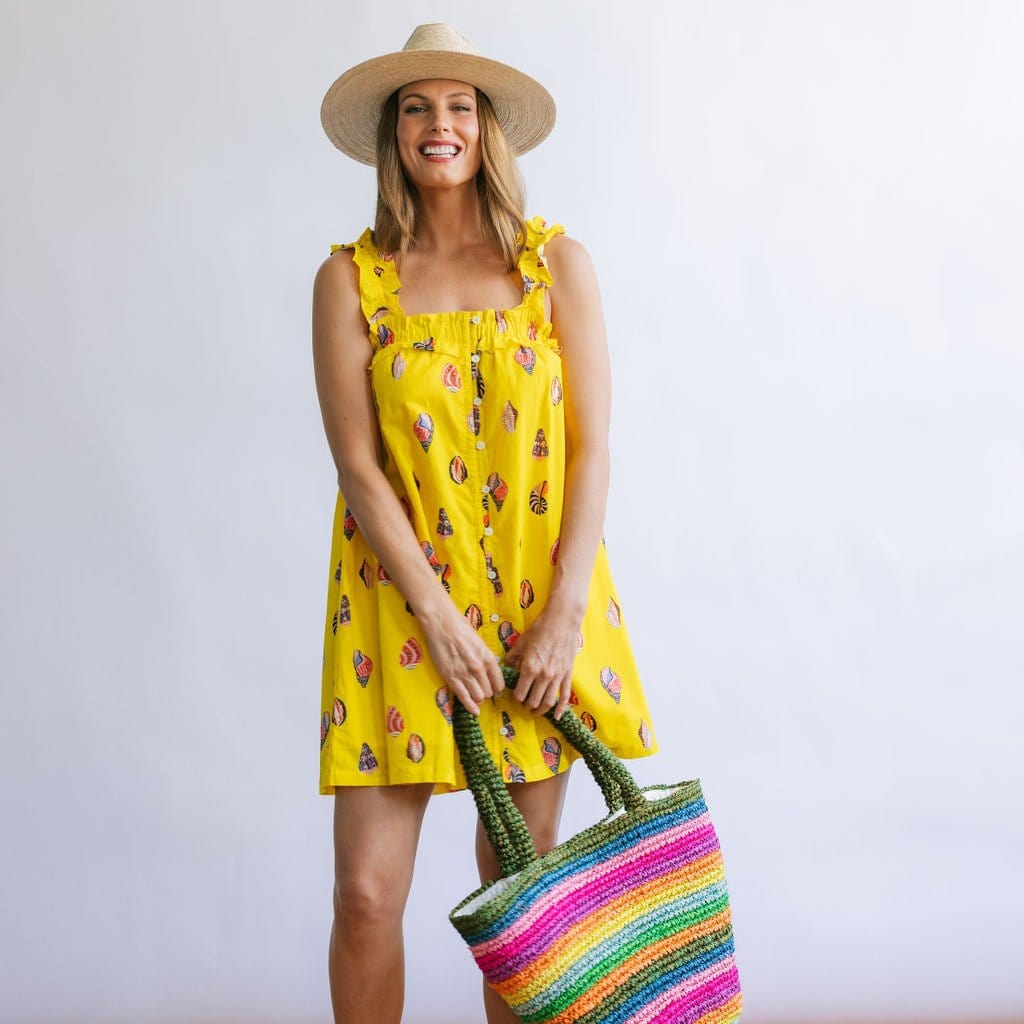 A woman wearing a yellow sleeveless mini dress with a seashell print, paired with a wide-brimmed hat and holding a multicolored striped tote bag.