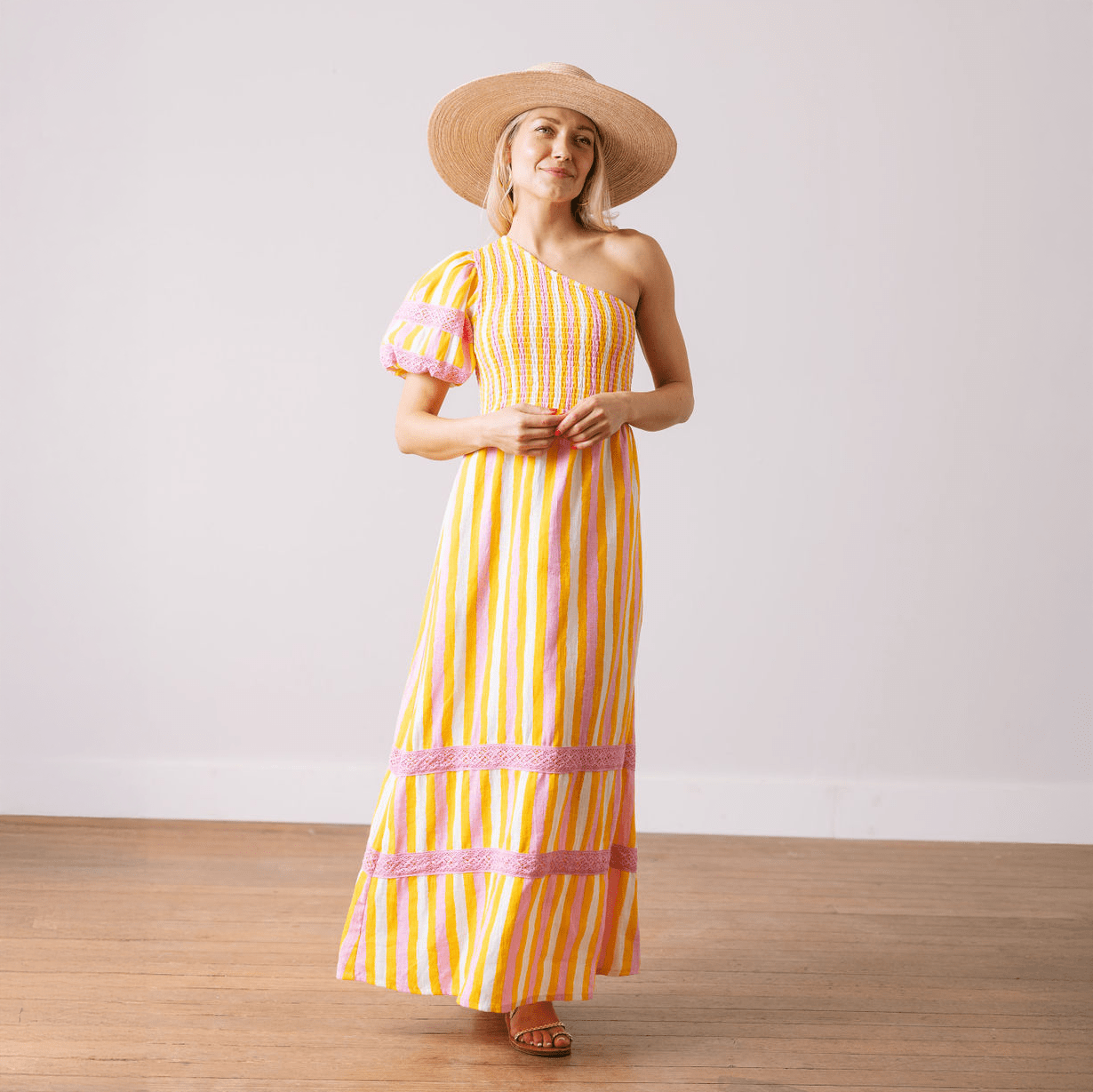 A model wearing a striped asymmetrical one-shoulder linen dress with a voluminous puff sleeve and a wide-brimmed hat.