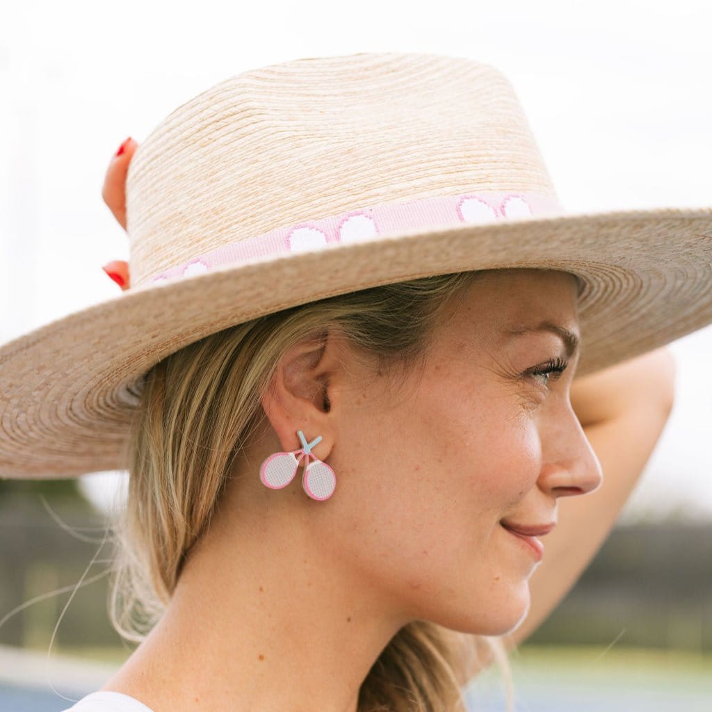A person wearing a straw hat and tennis doubles stud earrings with pink and turquoise handles.