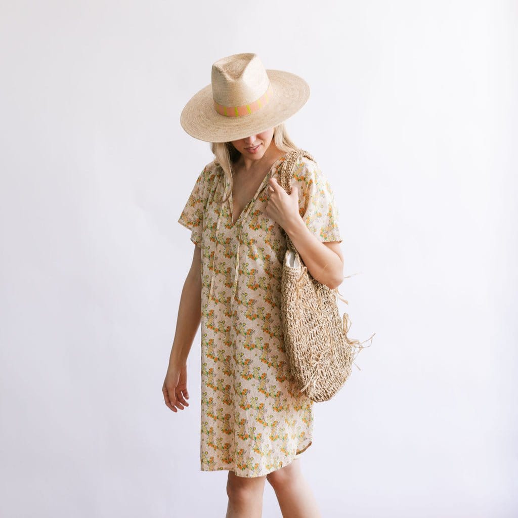 A female model wearing a floral print cover-up dress with short sleeves, paired with a wide-brimmed hat and a straw tote bag.