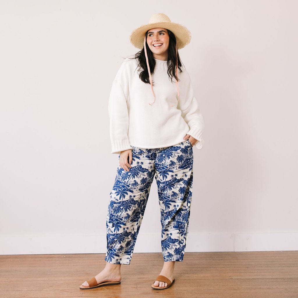Woman wearing a white sweater and blue floral pants with a straw hat against a plain background
