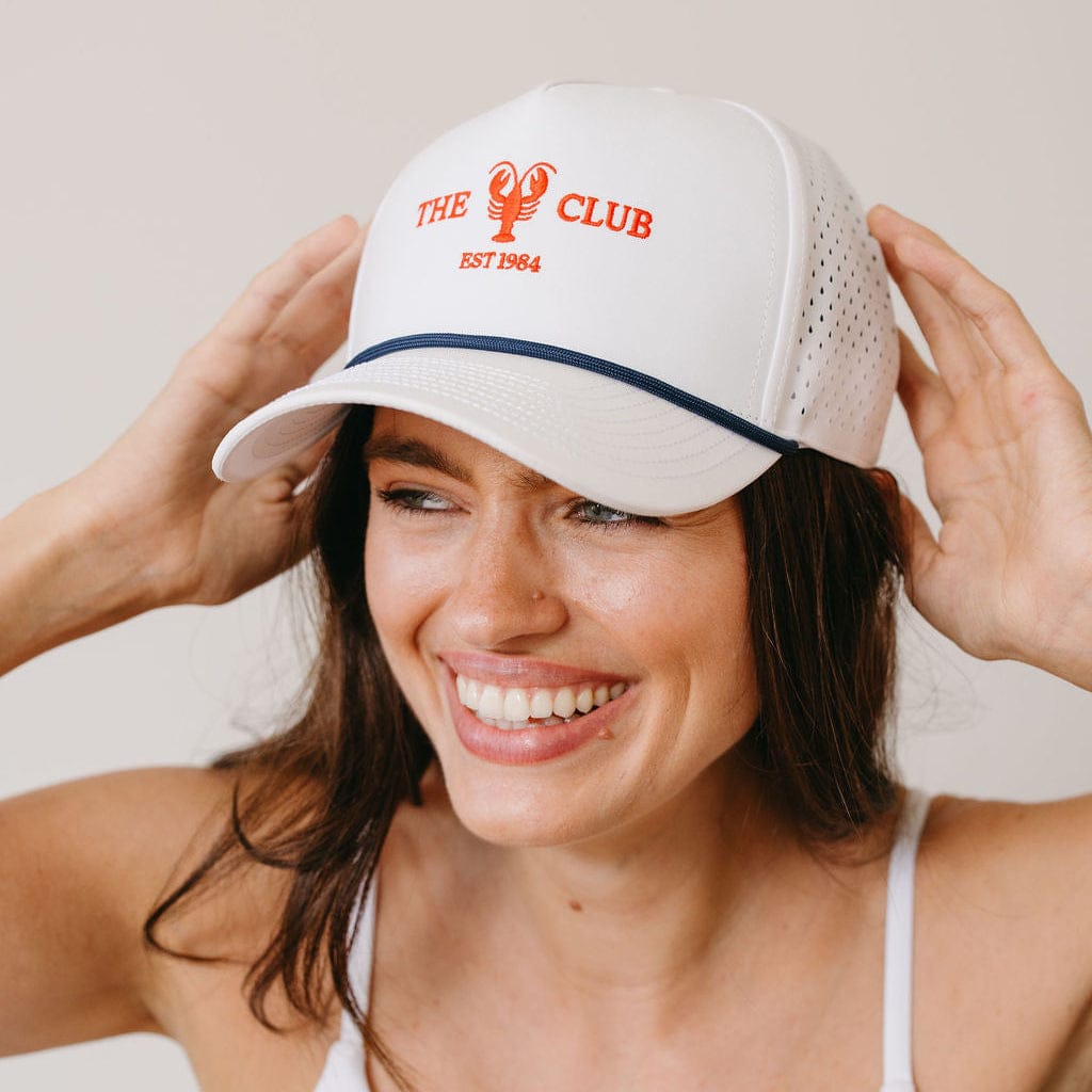 Woman wearing a white cap with 'The Club' logo, smiling against a plain background