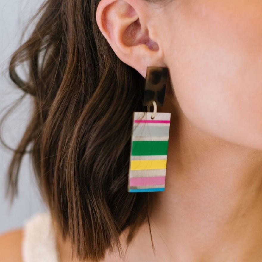 A close-up image of a person wearing dangle earrings with horizontal stripes of various colors and topped with a tortoise shell design.
